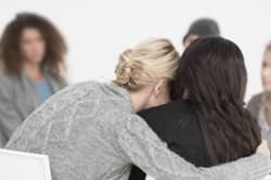 From behind, two women hug each other while sitting in recovery group